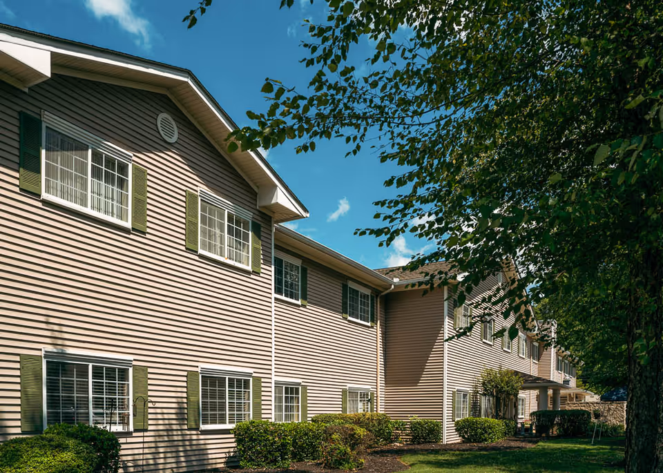 Exterior view of a two-story residential building with beige siding and green shutters, surrounded by bushes and trees under a blue sky with some clouds.