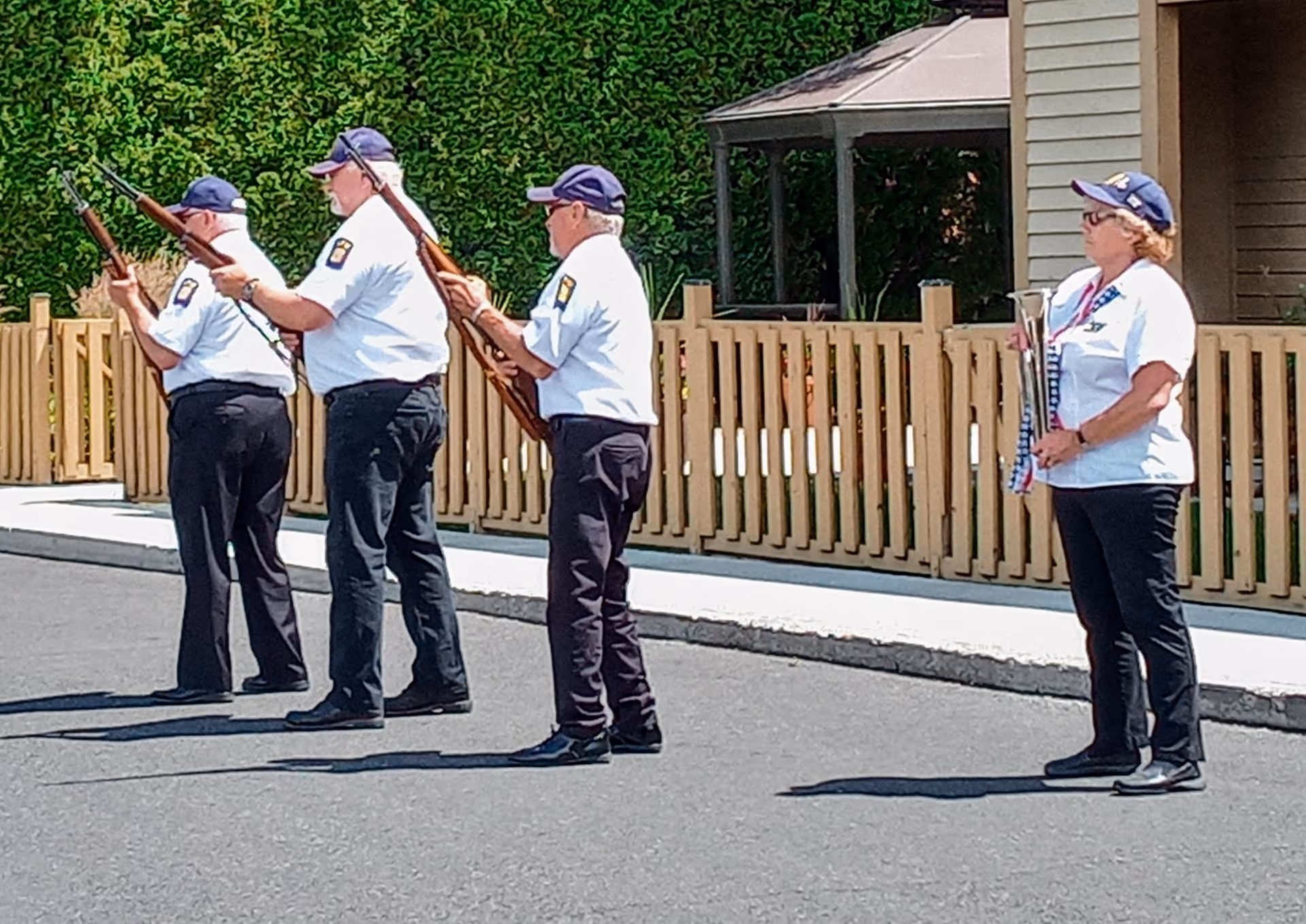Four people dressed in white shirts, black pants, and navy blue caps stand in a line outdoors on a paved surface. Three of them hold rifles pointed upwards, while the fourth person holds a folded American flag. Behind them is a wooden fence and greenery.