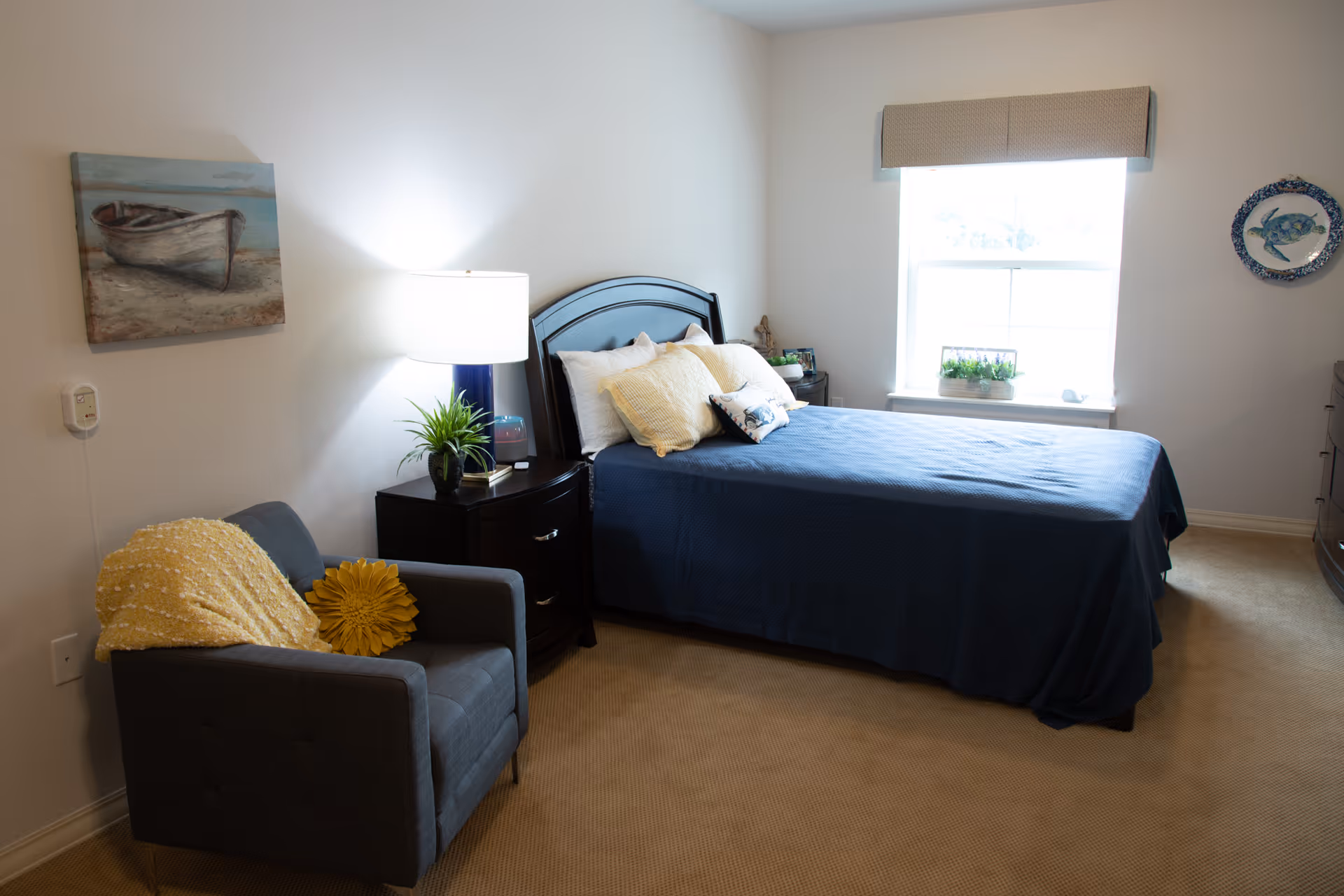 Neat bedroom with a bed covered in blue bedding, a nightstand with a lamp, an armchair with a yellow throw, and a window letting in natural light.