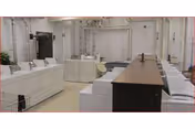 Large institutional laundry room with rows of white washers and dryers, folding tables, and overhead pipes.