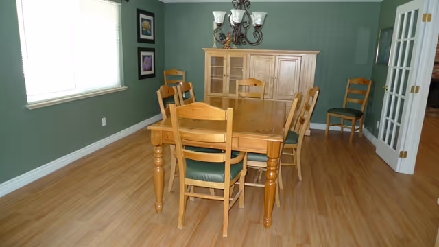 A dining room with a wooden dining table and six wooden chairs with green cushions. There is a wooden cabinet against a green wall, two framed pictures on the left wall, a window with blinds, and a chandelier hanging above the table. The floor is light wood, and there is a white door with glass panels on the right side.