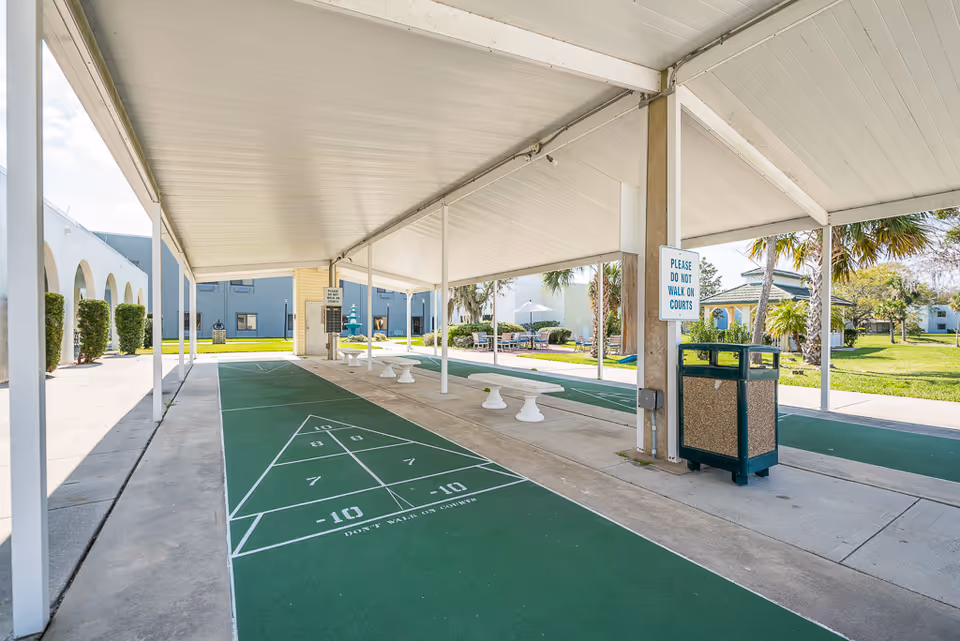 Covered outdoor shuffleboard courts with seating, a trash receptacle, and surrounding apartment buildings.