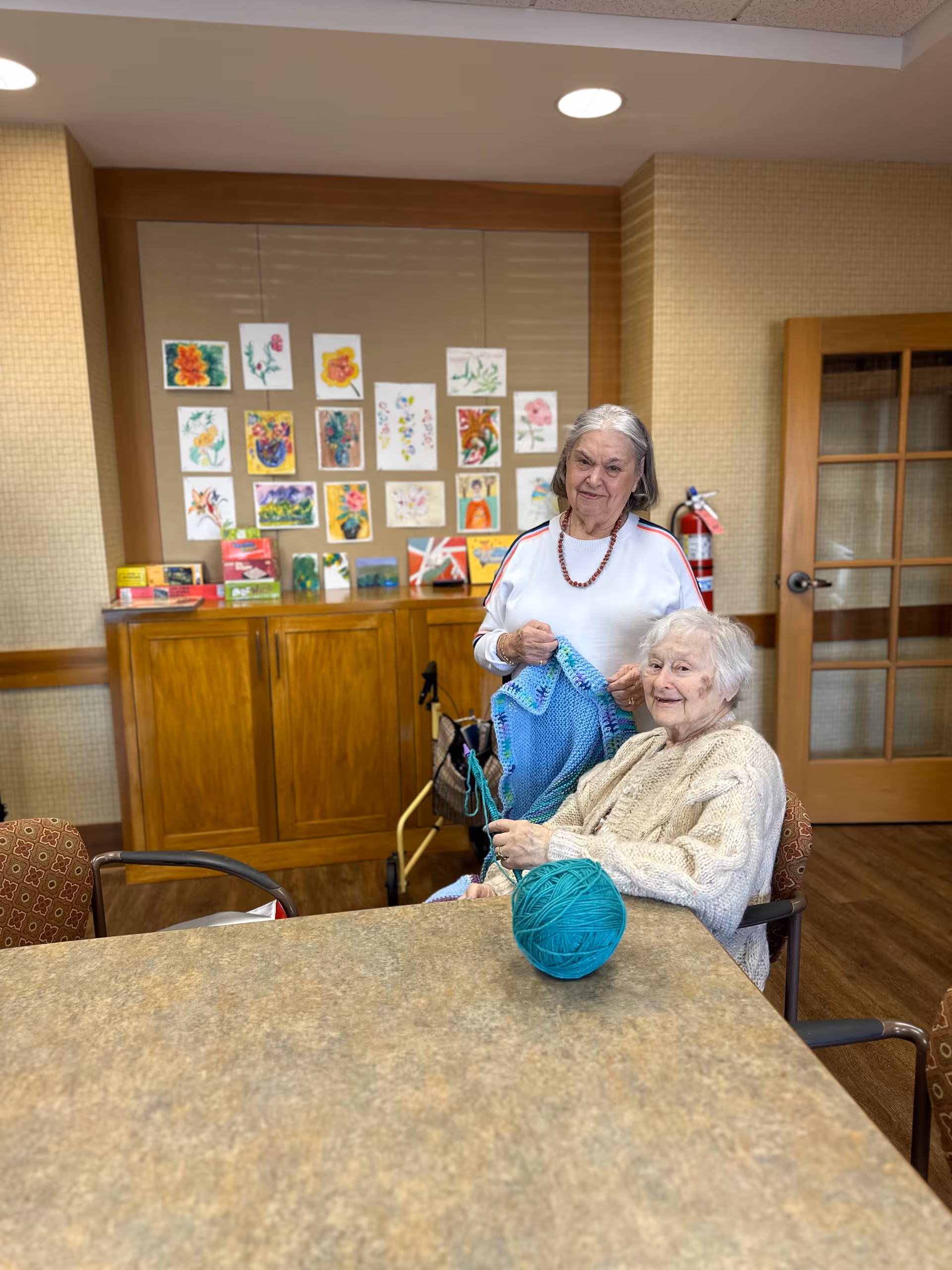 Two elderly women in a room with wooden furniture and a bulletin board displaying colorful floral artwork. One woman is sitting and knitting with blue yarn, while the other stands beside her holding a knitted piece. The room has a warm, inviting atmosphere with a fire extinguisher mounted on the wall and a door with glass panels in the background.