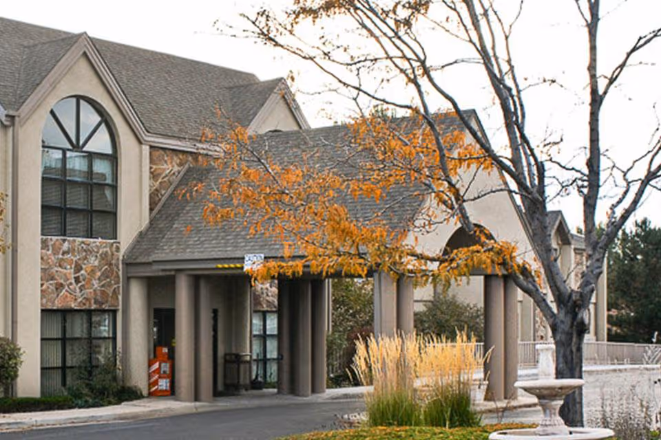 Exterior view of the Life Care Center of Longmont building entrance with a covered driveway, stone and stucco facade, large windows, and autumn trees with orange leaves in the foreground.