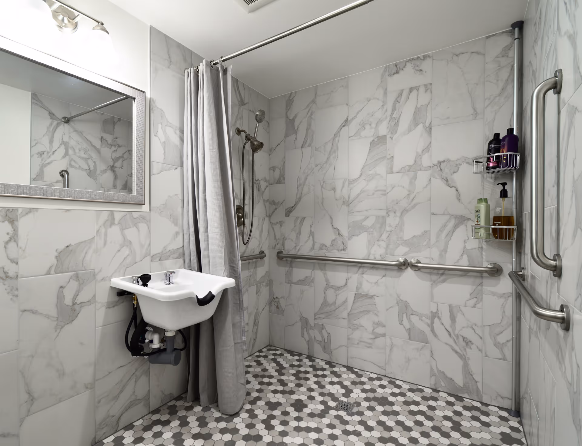 Accessible bathroom with marble-patterned wall tiles and hexagonal floor tiles. The room features a wall-mounted sink with exposed plumbing, a large mirror above it, a shower area with a curtain, a handheld showerhead, and multiple stainless steel grab bars for support. A shower caddy holds various toiletries.