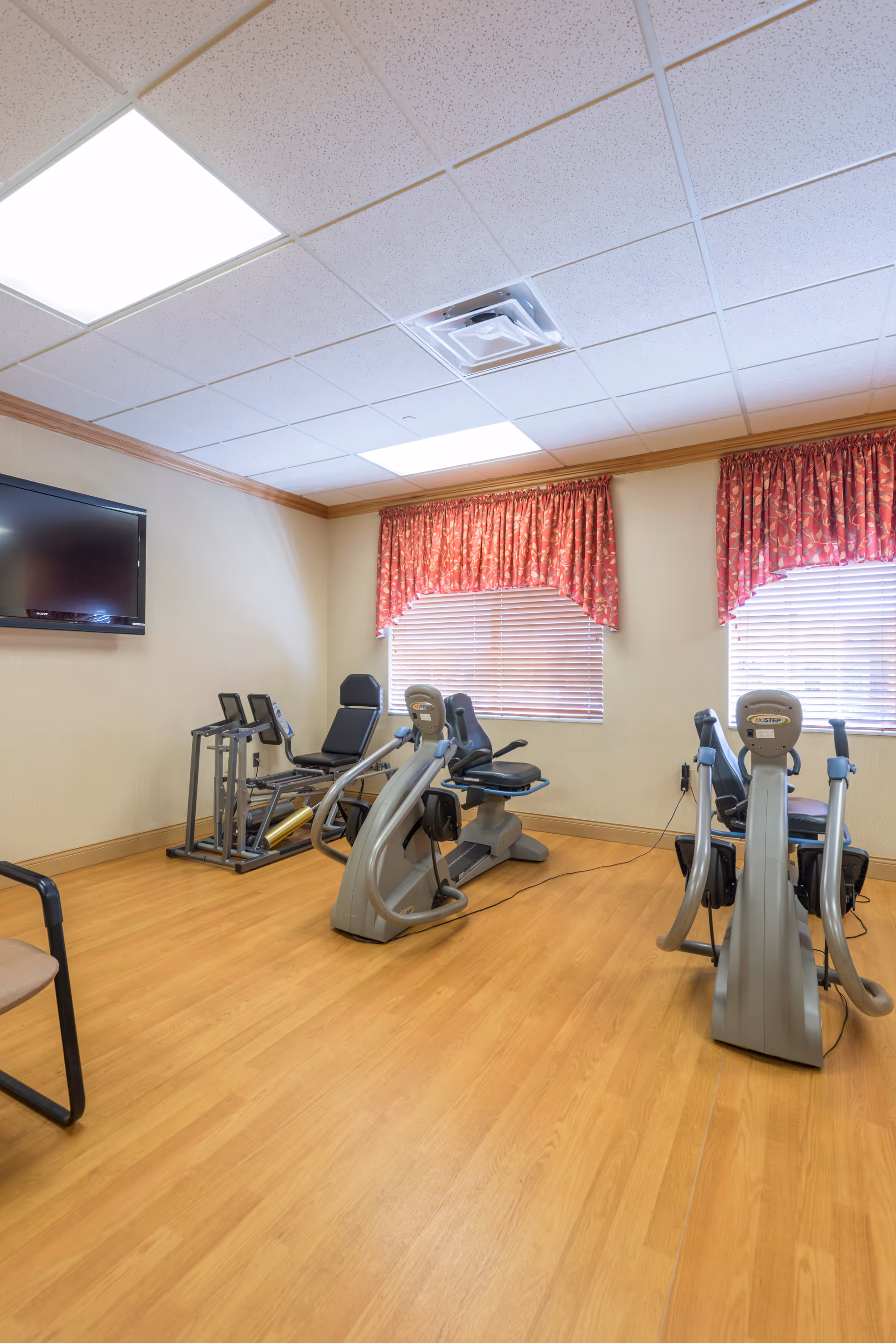 A small exercise room with wooden flooring, two recumbent exercise bikes, a stepper machine, a chair, and a wall-mounted flat screen TV. The room has two windows with red patterned curtains and blinds, and a white drop ceiling with fluorescent lights.