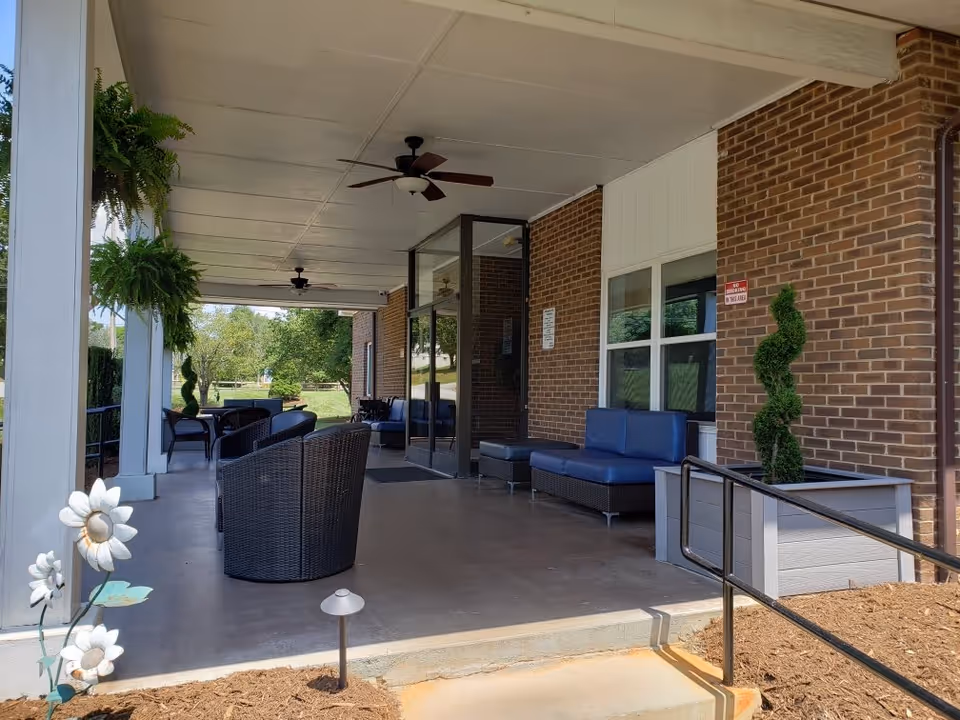 Covered outdoor patio area with wicker chairs and blue cushioned seating along a brick wall. Ceiling fans are mounted on the white ceiling. There are potted plants and decorative flowers near the entrance, with a ramp and steps leading up to the patio. Green trees and grass are visible in the background.