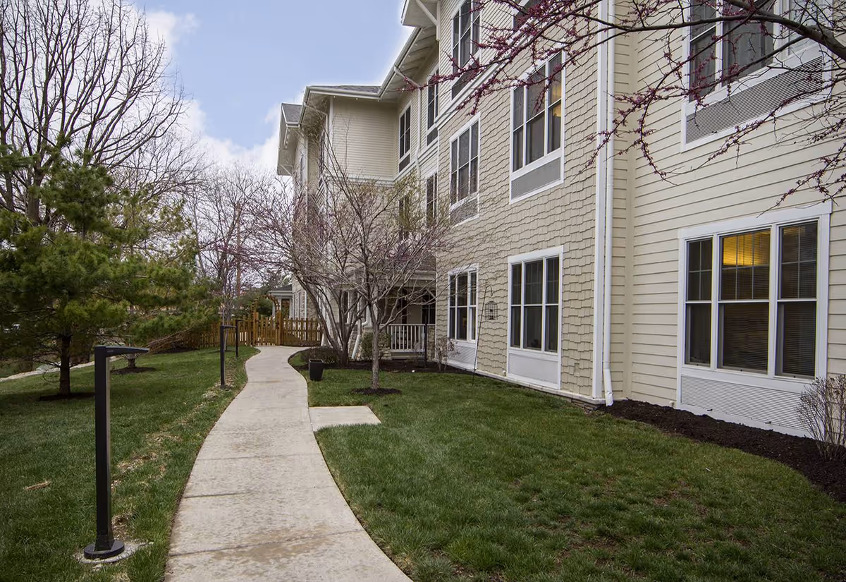 A curved concrete walkway passes a multi-story beige senior living building with windows, lawn, and trees.