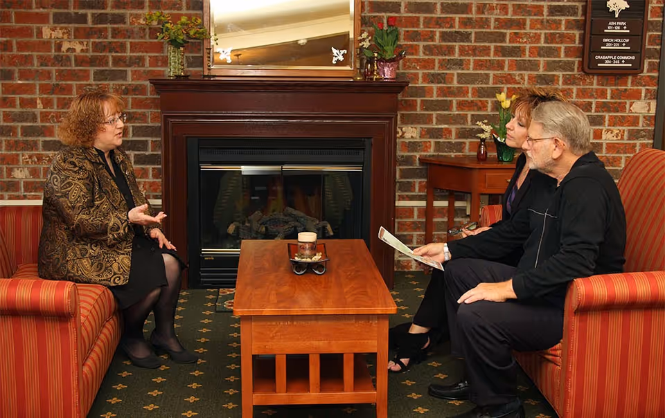 Three people sit on sofas in a cozy living room area with a brick fireplace and a wooden coffee table.