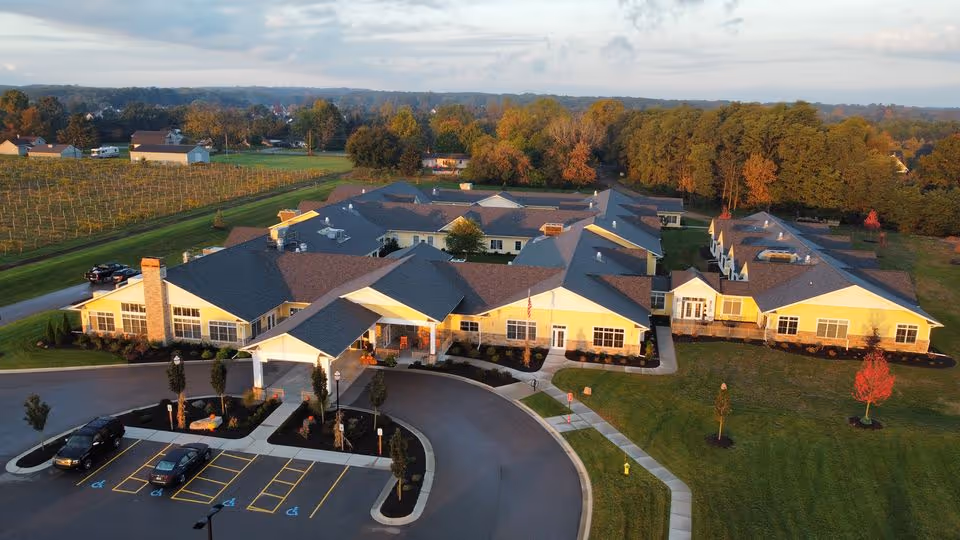 Aerial view of Vineyard Assisted Living Community, showing a large single-story building with multiple wings surrounded by green lawns, trees, and a parking lot with several cars. The building has a covered entrance and is set in a rural area with fields and wooded areas in the background.