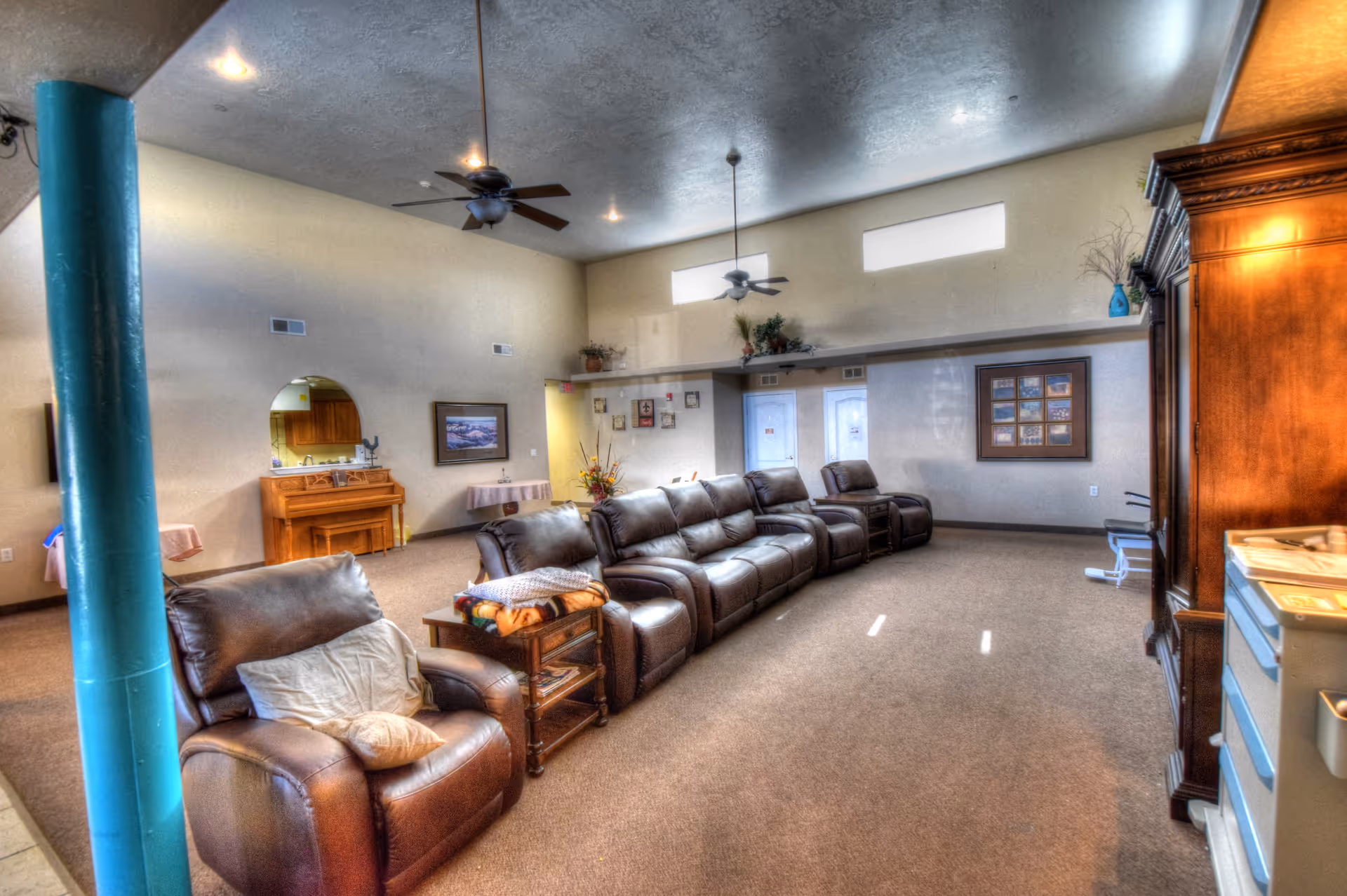 Spacious communal living room with a row of brown leather recliner sofas, ceiling fans, and an entertainment cabinet under high windows.