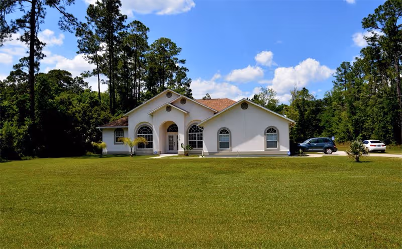A single-story white building with arched windows and a central entrance, surrounded by a large grassy lawn and tall trees under a blue sky with scattered clouds. Two cars are parked on the right side of the building.