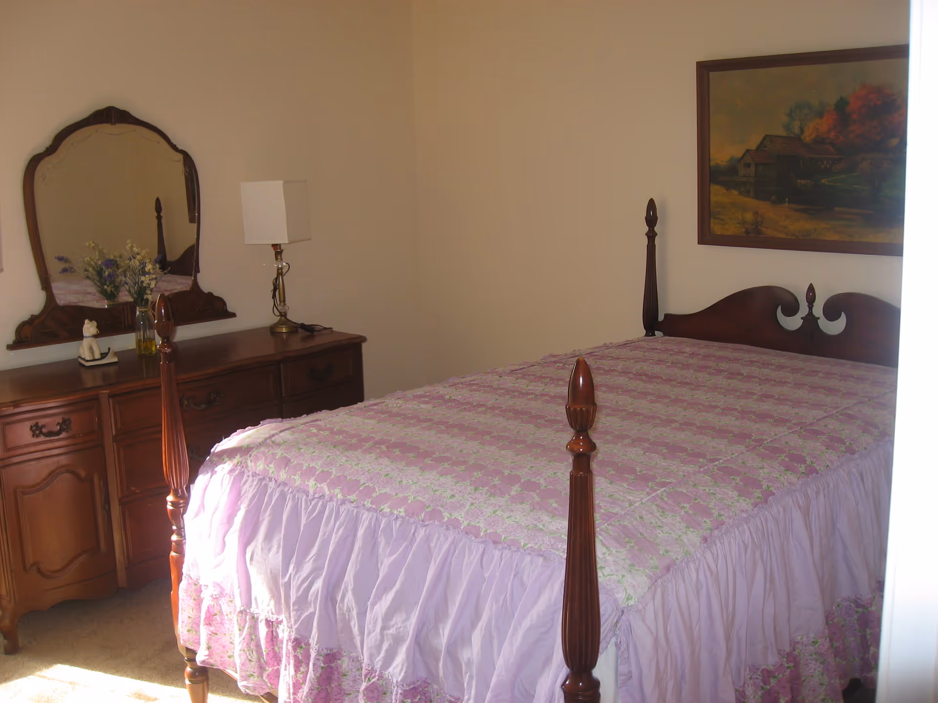 A bedroom with a wooden four-poster bed covered with a pink and white quilt. To the left is a wooden dresser with an ornate mirror, a table lamp, a vase with flowers, and a small decorative cat figurine. A framed painting of a rustic landscape hangs on the wall above the bed.
