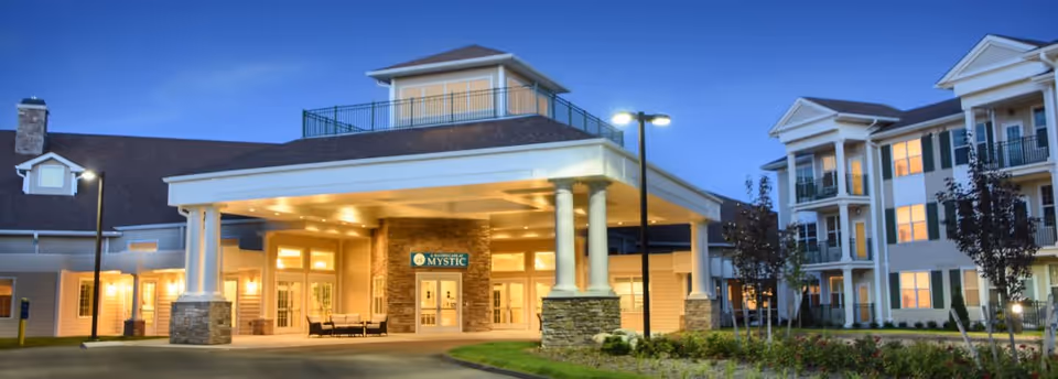 Exterior view of the Masonicare At Mystic senior living facility at dusk, showing the main entrance with a covered drop-off area supported by columns, well-lit windows, and a multi-story building with balconies and landscaping.