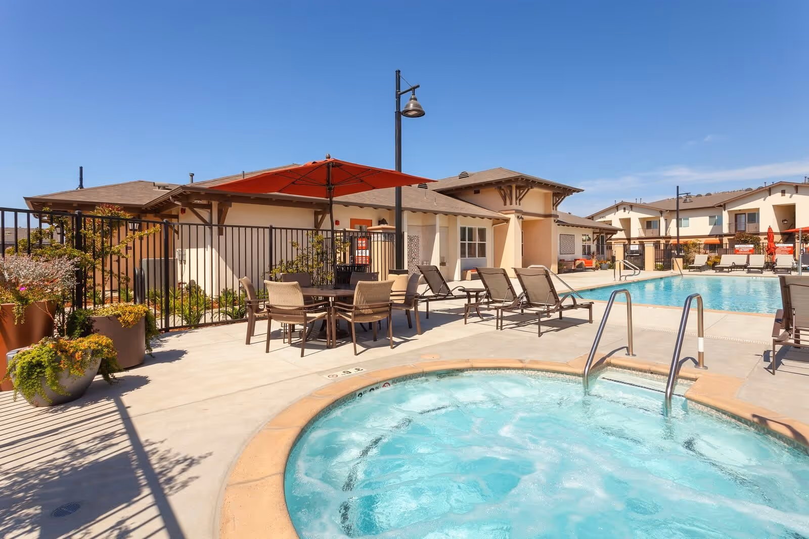 Outdoor pool area at Villa del Sol featuring a hot tub in the foreground, a swimming pool in the background, several lounge chairs, a table with chairs under a red umbrella, potted plants, and residential-style buildings under a clear blue sky.
