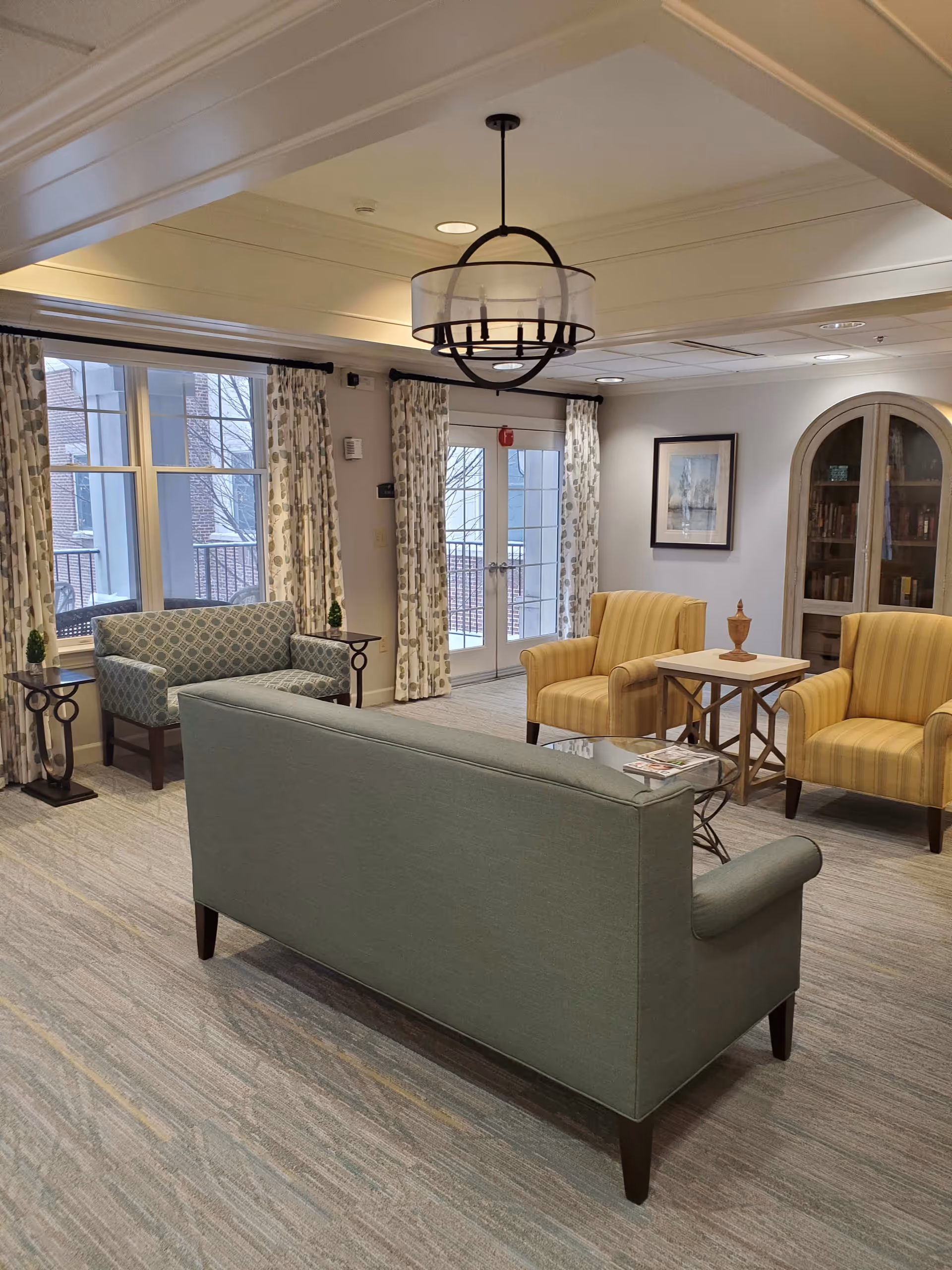 A cozy sitting area in a senior living facility with a green sofa, two yellow armchairs, and a patterned loveseat. The room has large windows with patterned curtains, a glass door leading outside, a chandelier hanging from the ceiling, a framed picture on the wall, and a wooden cabinet with glass doors containing books.