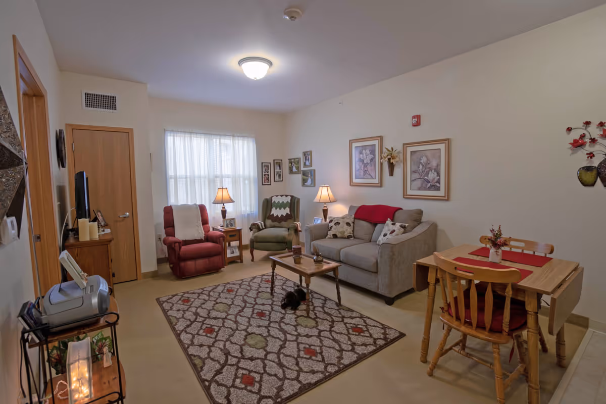 A cozy living room in a senior living facility with a beige sofa, two armchairs (one red and one green), a wooden coffee table on a patterned rug, two table lamps, framed floral artwork on the walls, and a small wooden dining table with two chairs and red placemats.