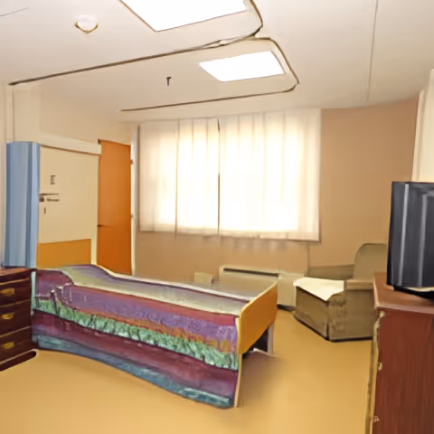 Single-occupancy room with a bed covered by a colorful blanket, an armchair, dresser with a TV, and a curtained window.