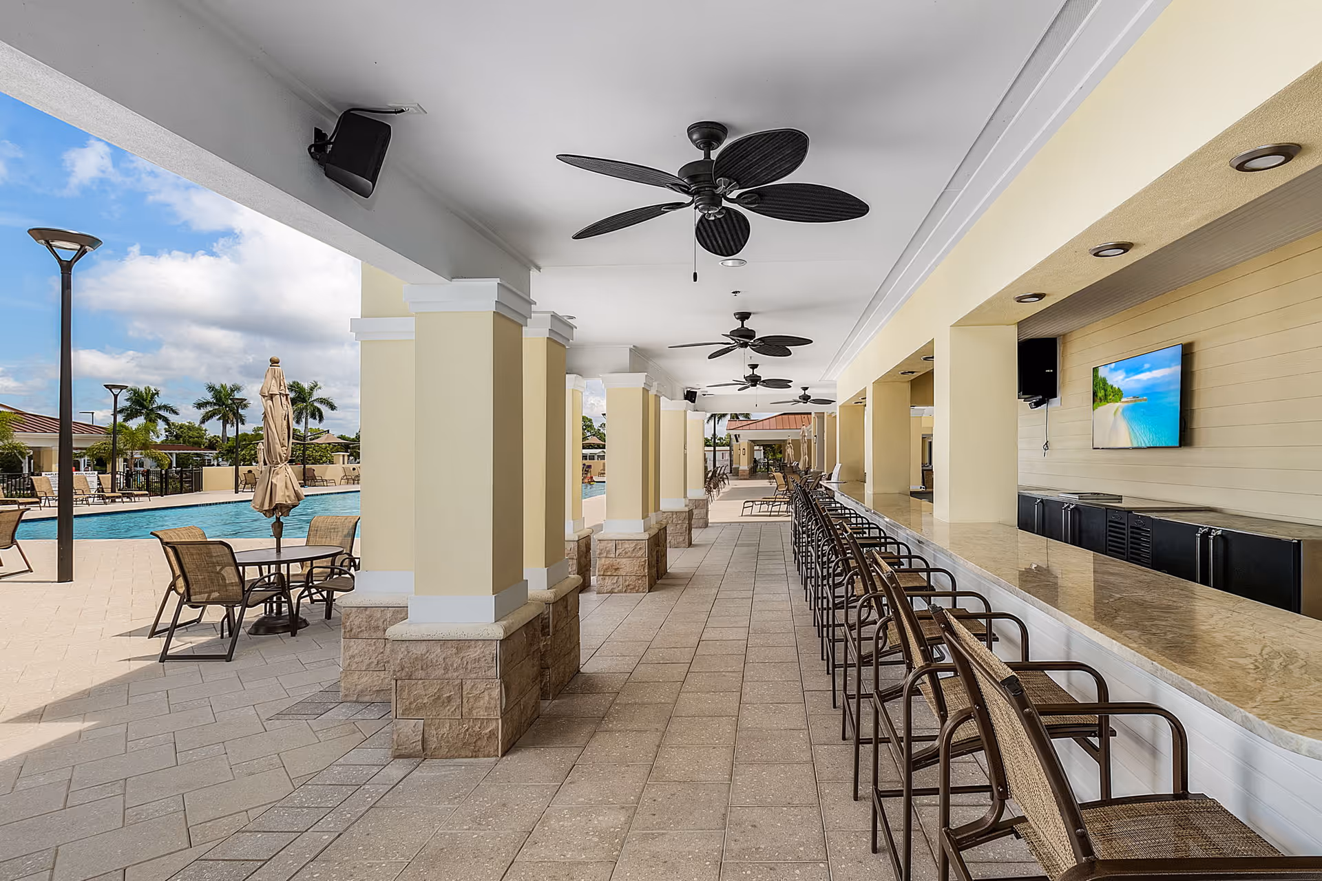 Outdoor covered patio area with ceiling fans and a long counter with bar stools facing a mounted TV. Adjacent to the patio is a swimming pool with tables, chairs, and umbrellas. Palm trees and a partly cloudy sky are visible in the background.