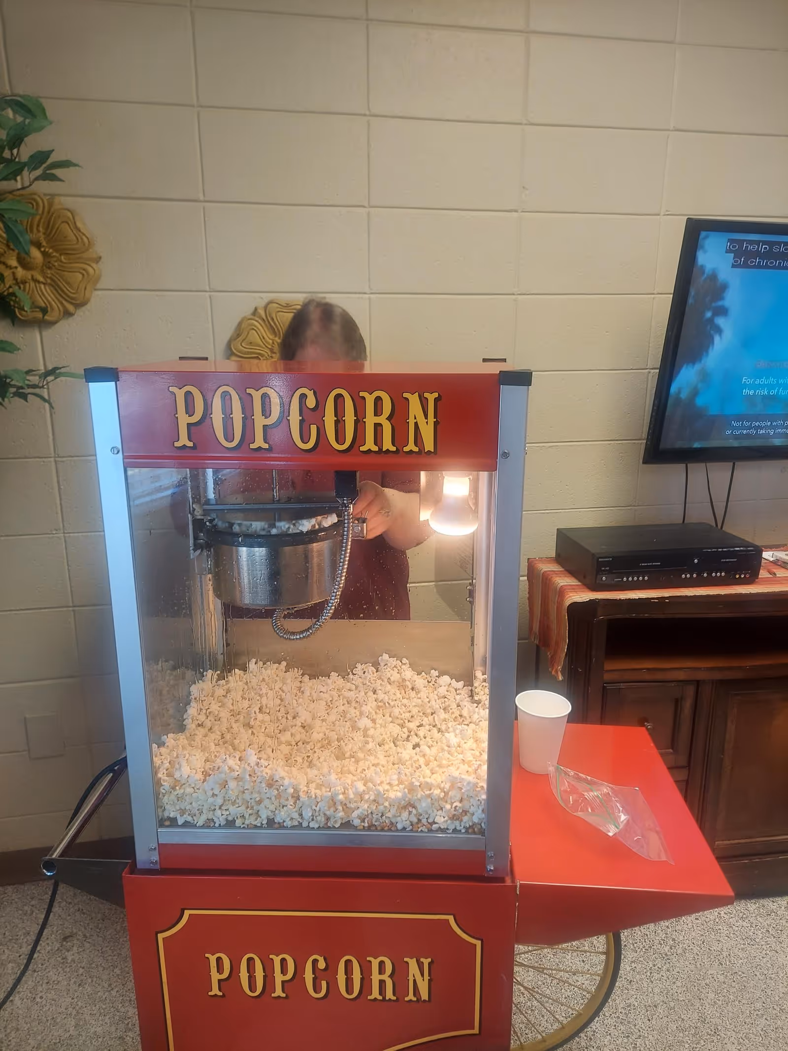 Red vintage popcorn machine on a cart filled with popcorn in an interior common room, with a person behind it and a TV on a cabinet to the right.