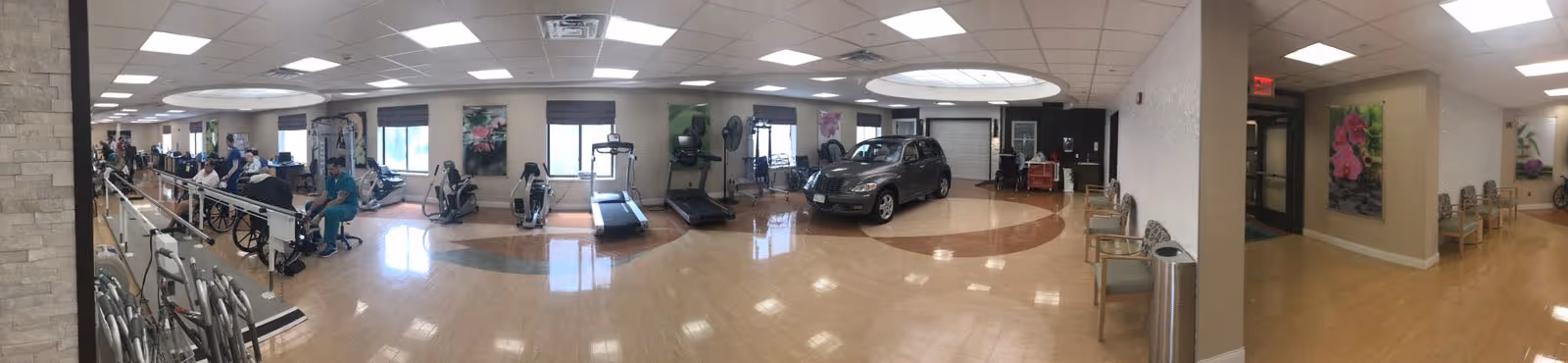 A spacious rehabilitation or physical therapy room with exercise equipment including stationary bikes, treadmills, and a car used for therapy. Several people are seated, some in wheelchairs, and a few staff members are present. The room has large windows allowing natural light, light-colored walls, and a polished wooden floor. Chairs are arranged along the walls, and there are decorative flower paintings on the walls.