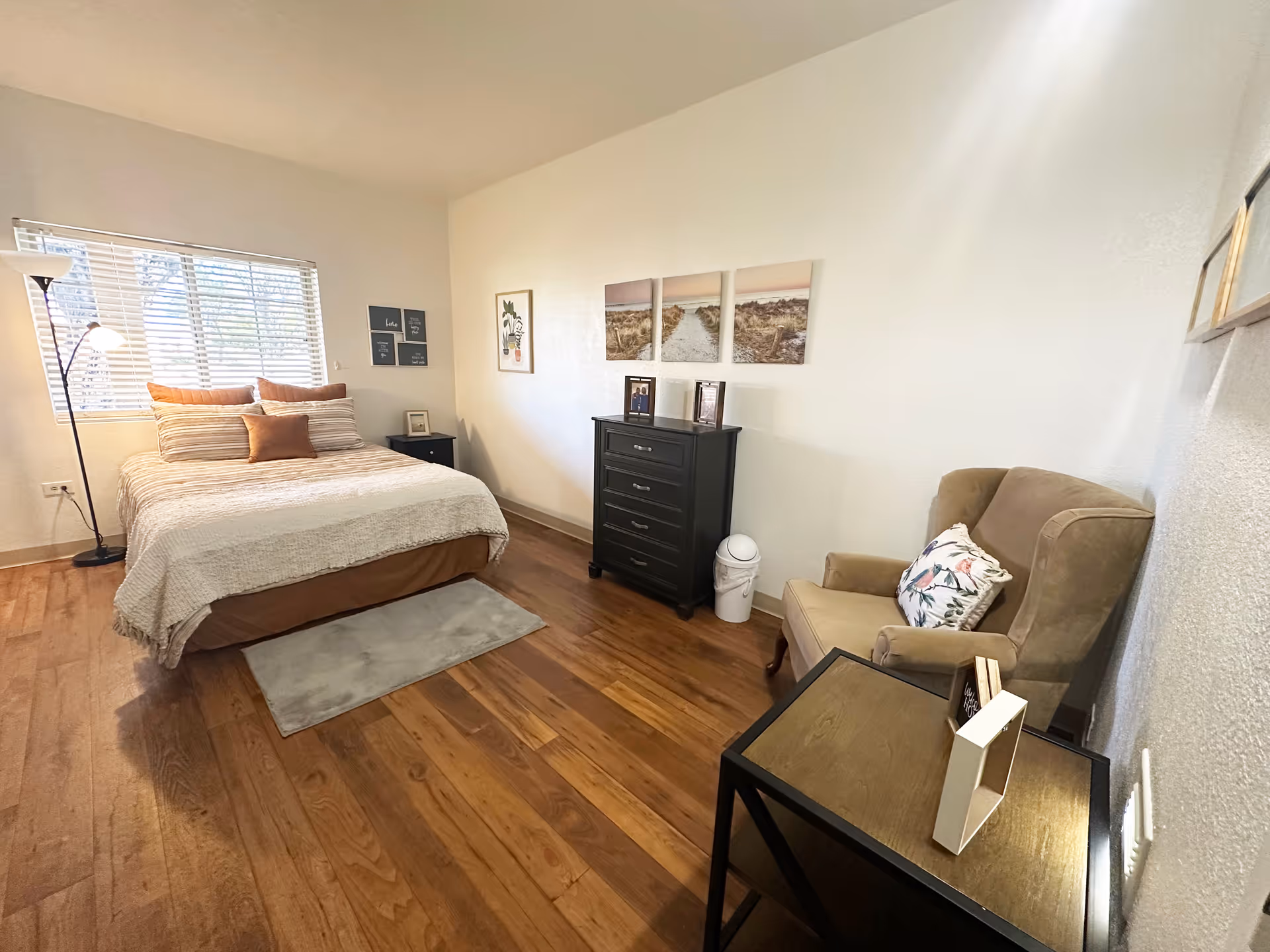 A bright furnished bedroom with a made bed, dresser, armchair and side tables on hardwood floors.