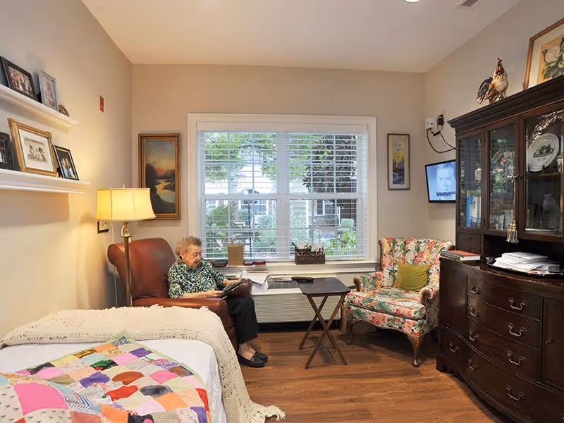 A cozy room in a senior living facility with a bed covered by a colorful quilt and a white crocheted blanket. An elderly woman is sitting in a brown leather armchair reading a book. Next to her is a floral upholstered armchair and a small wooden table. A large window with white blinds lets in natural light, and a dark wooden cabinet with glass doors displays plates and other items. The walls are decorated with framed pictures and a lamp provides warm lighting.