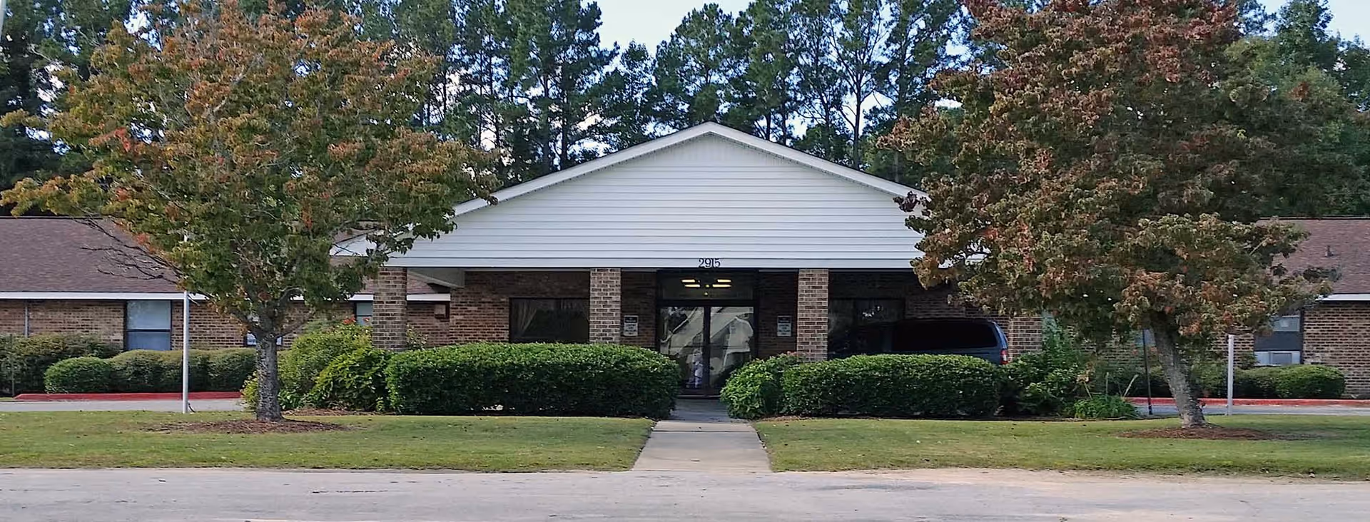 Front exterior view of a single-story brick building with a white gabled roof entrance, surrounded by green bushes and trees, with a paved walkway leading to the entrance.