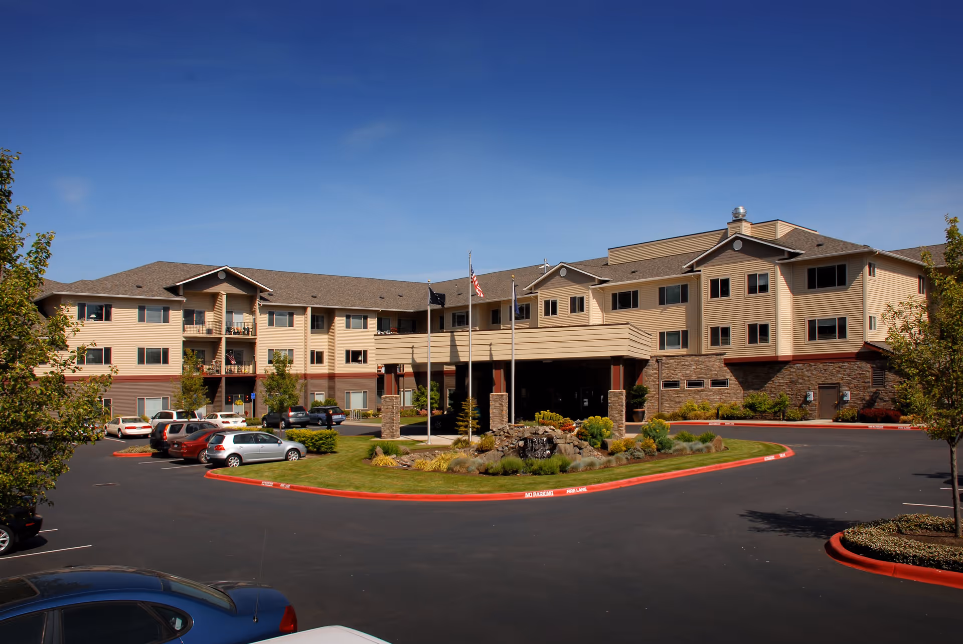 Exterior view of a three-story senior living community building with beige and brown siding, multiple windows, and a covered entrance. There are three flagpoles with flags in front of the entrance, a landscaped area with rocks and plants, and a parking lot with several cars parked. The sky is clear and blue.