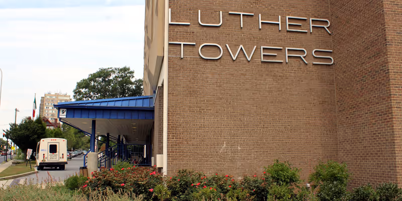 Exterior view of a brick building with the sign 'Luther Towers' mounted on the wall. There is a blue awning over the entrance and some bushes with flowers in front of the building. A white bus is parked on the street nearby.