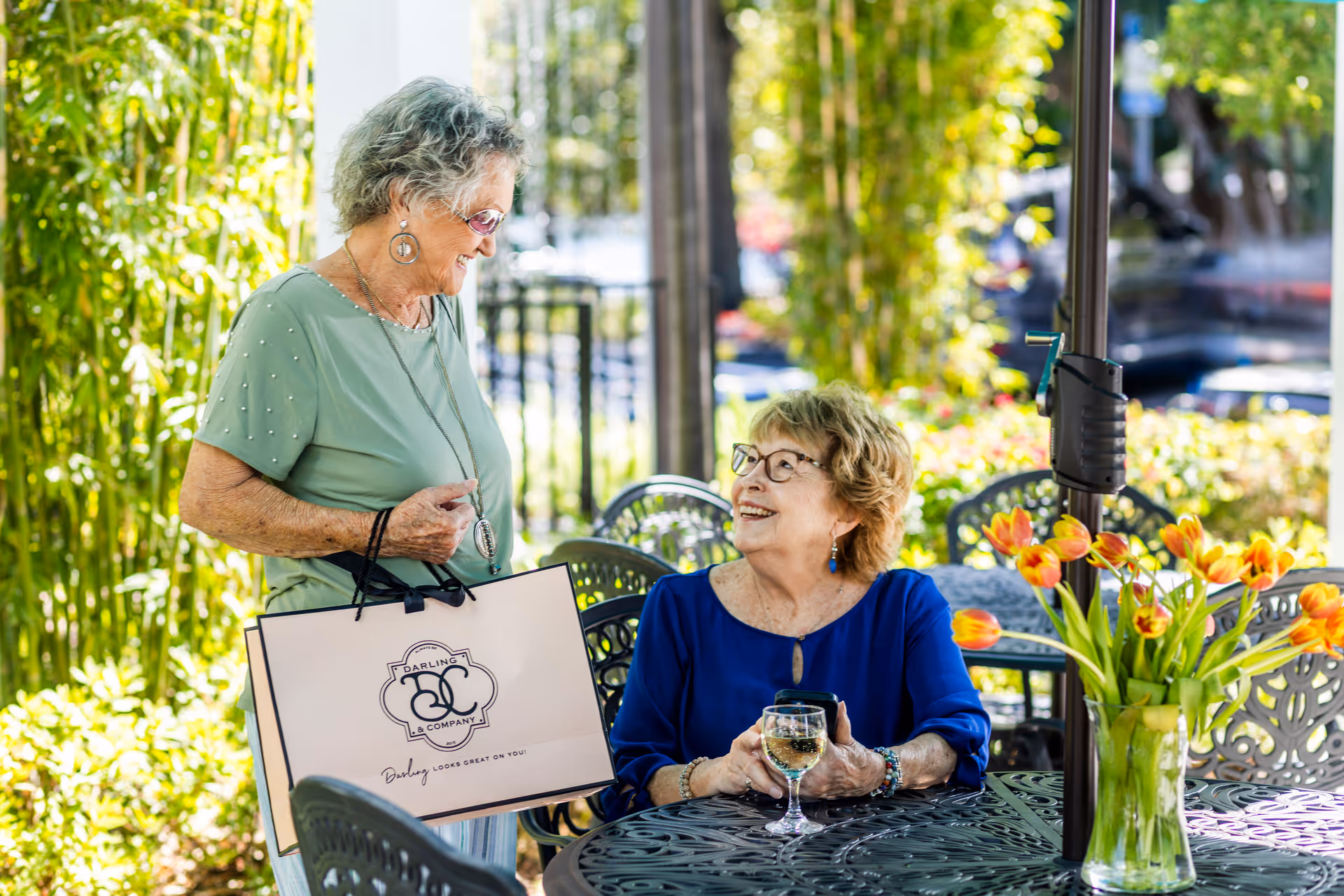 Two elderly women enjoying a sunny day outdoors at a patio table. One woman is standing holding a shopping bag and smiling, while the other is seated holding a glass of white wine and looking up at her with a smile. The table has a vase with orange tulips, and the background features green plants and trees.