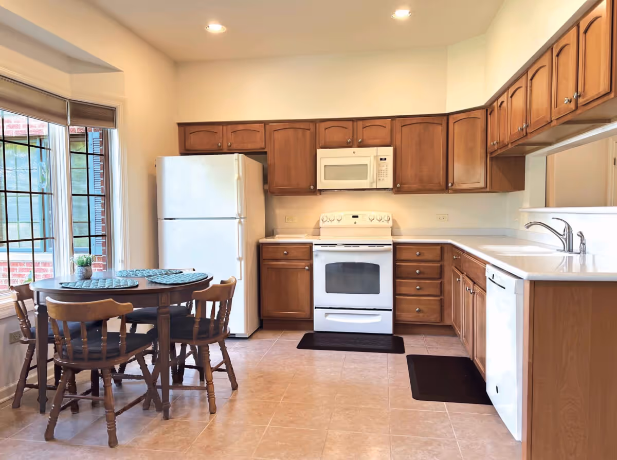A bright kitchen with wooden cabinets, a white refrigerator, white stove with oven, white microwave, and white dishwasher. There is a round wooden dining table with four wooden chairs near a large window with blinds. The floor is tiled and the walls are painted light beige.