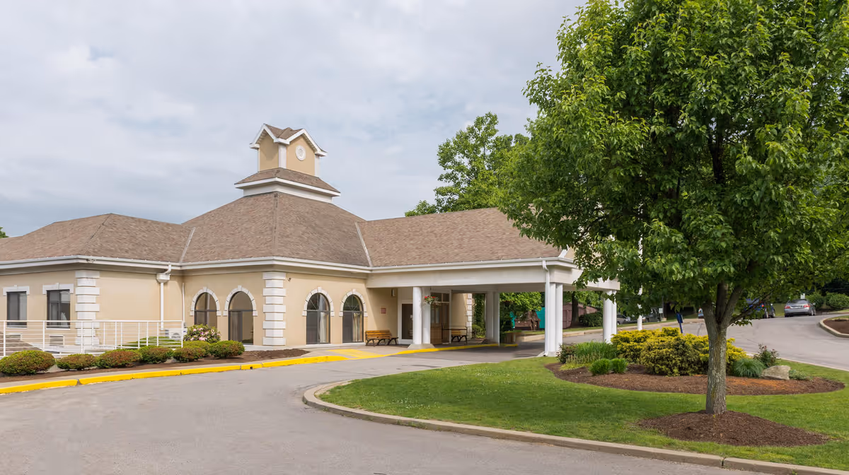 Front entrance of Highland Hills Post Acute featuring a porte-cochère, benches, and landscaped grounds with a large tree.