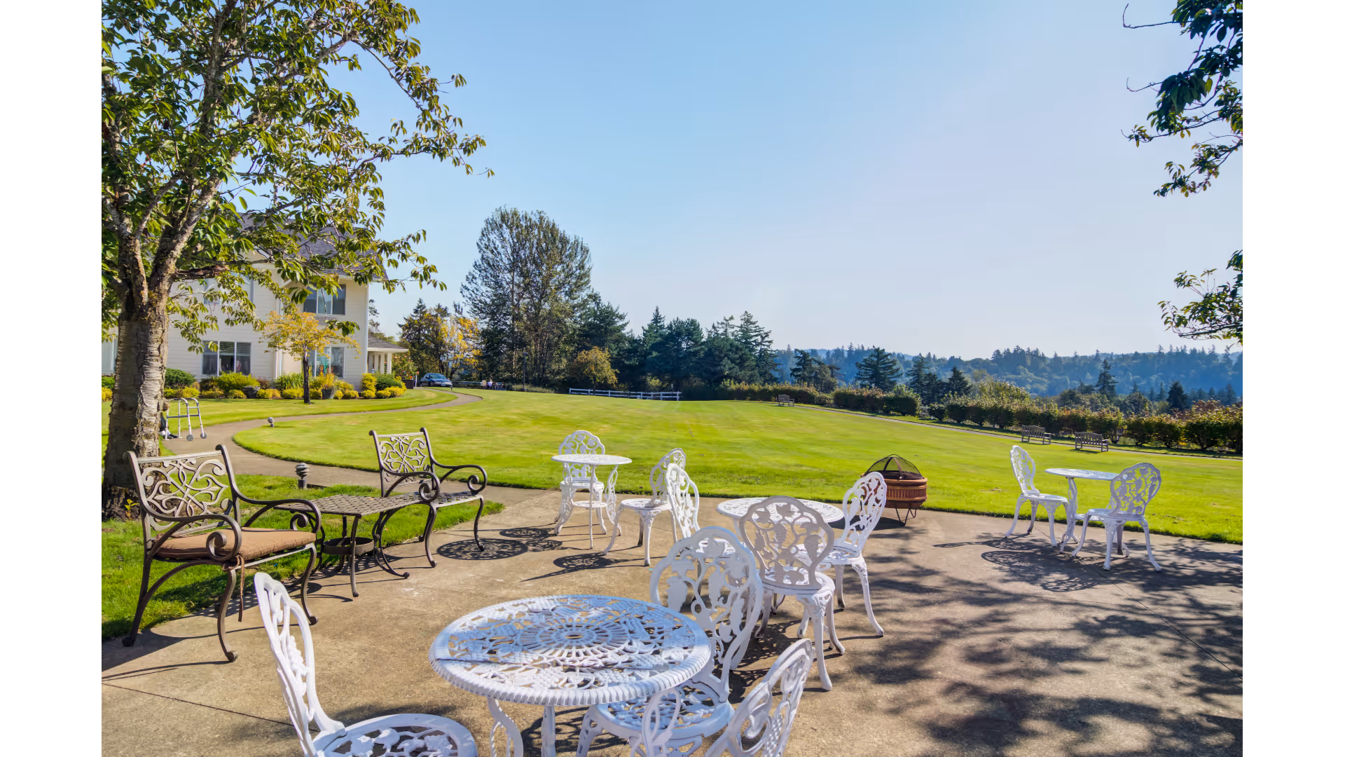 Outdoor patio area with several white metal tables and chairs arranged on a concrete surface, surrounded by green grass and trees under a clear blue sky. A building is partially visible on the left side with a walker nearby.