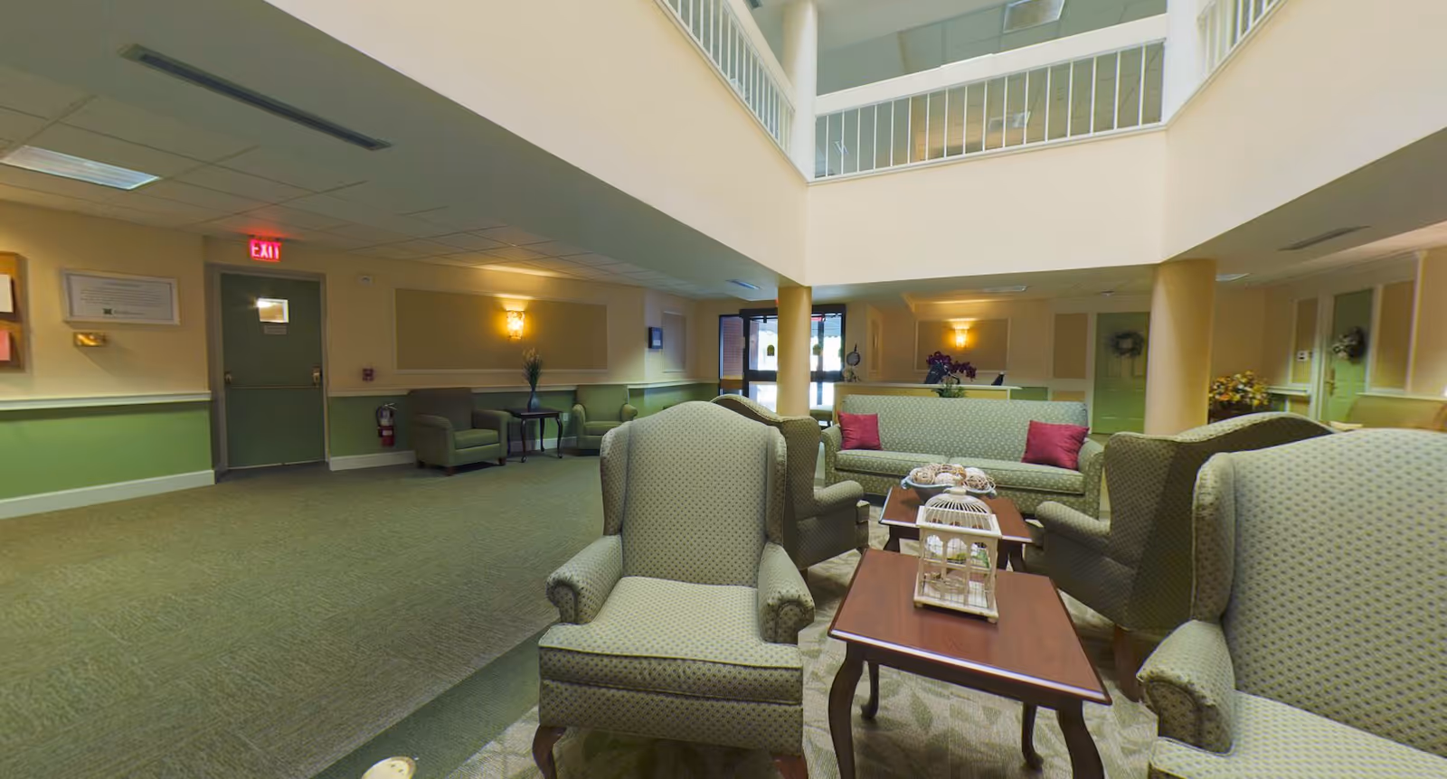 A spacious and well-lit common area in a senior living facility with green upholstered armchairs and sofas arranged around wooden coffee tables. The room features a high ceiling with a balcony railing above, soft wall lighting, and green and beige walls. There are decorative items on the tables and a fire extinguisher mounted on the wall near a green door with an exit sign above it.