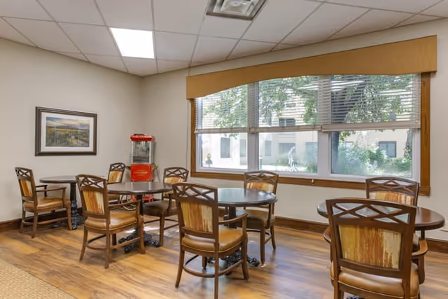 A small dining area with several round wooden tables and matching chairs with cushioned seats. There is a large window with blinds and a valance, letting in natural light and showing greenery outside. A red popcorn machine is placed in the corner near a framed landscape picture on the wall. The floor is wooden, and the ceiling has recessed lighting.