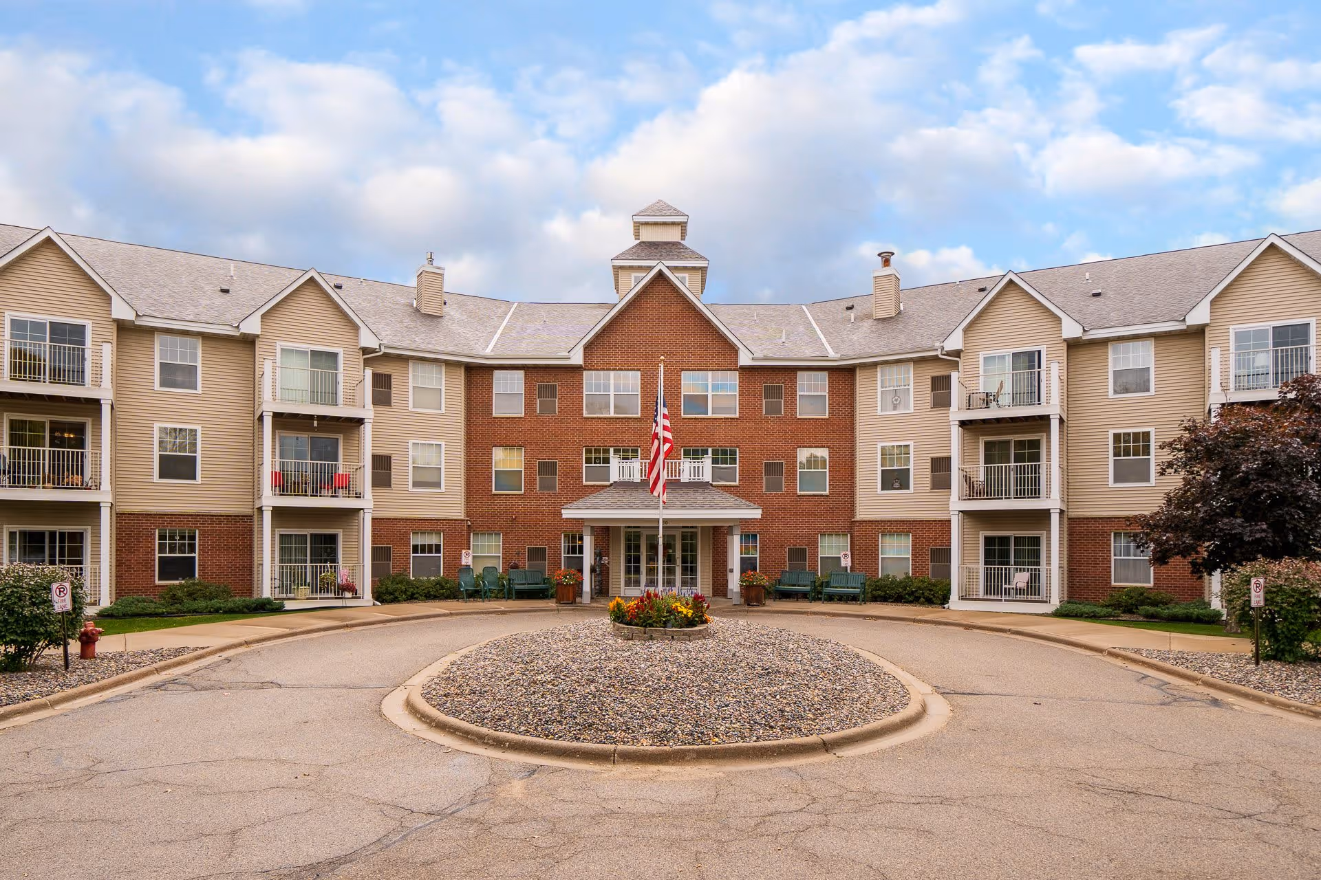 Front exterior view of a senior living facility with a circular driveway, an American flag in the center, and a three-story building with balconies and windows under a partly cloudy sky.