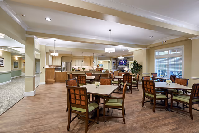 Spacious communal dining area with multiple square tables and chairs, a serving counter/kitchen in the background, and pendant lighting.