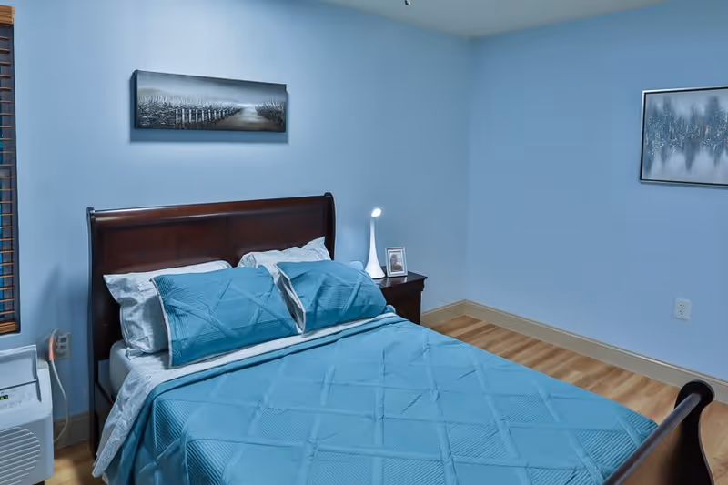 A neatly made bed with blue bedding and pillows in a bedroom with light blue walls. There is a wooden headboard, a nightstand with a small lamp and a framed photo, two paintings on the walls, and a window with wooden blinds.