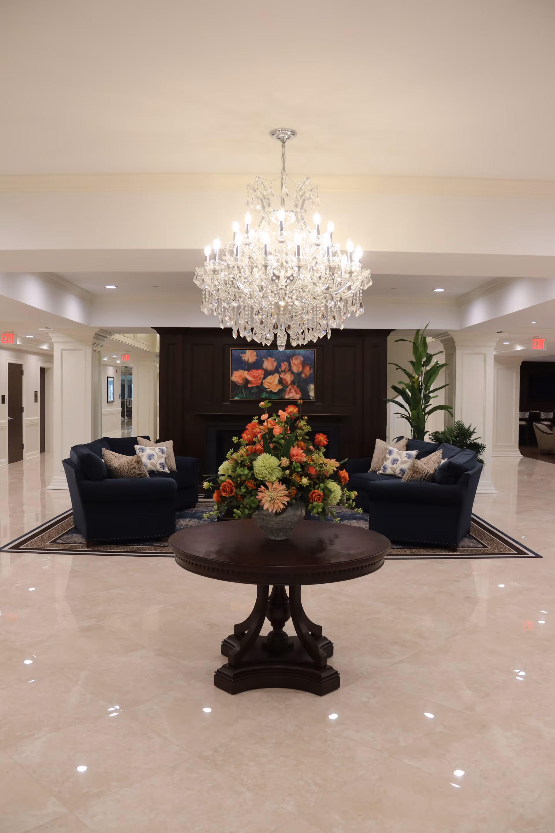 Elegant interior lobby with a large crystal chandelier, a round table topped with a floral arrangement, and a seating area with sofas.