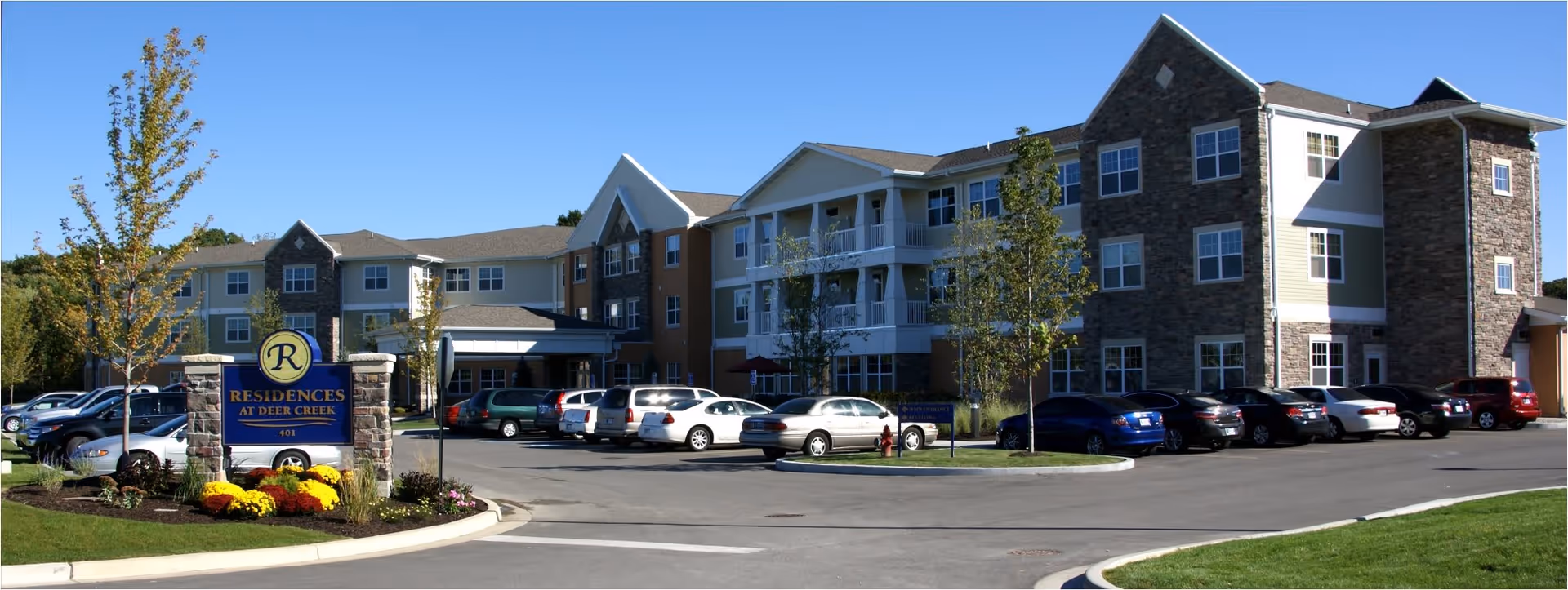 Exterior view of a multi-story senior living facility named Residences At Deer Creek with a parking lot in front, several cars parked, and landscaped greenery including trees and flowers around the entrance sign.