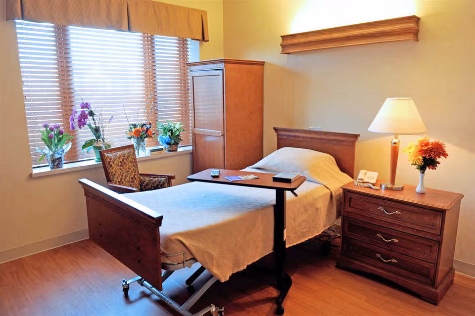 A nursing facility bedroom with a single bed covered in a beige blanket, a wooden nightstand with a lamp and a vase of flowers, a wooden cabinet, a patterned armchair, and a window with wooden blinds and several potted plants on the windowsill.