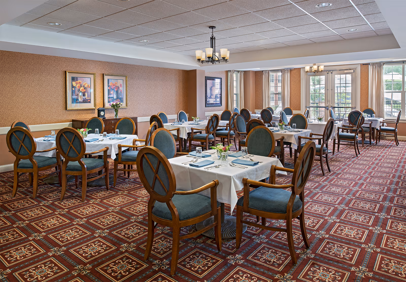 A dining room with multiple tables covered in white tablecloths, each set with glasses, napkins, and small flower arrangements. The room has patterned red carpet, wooden chairs with blue cushions, framed floral artwork on the walls, and large windows letting in natural light.