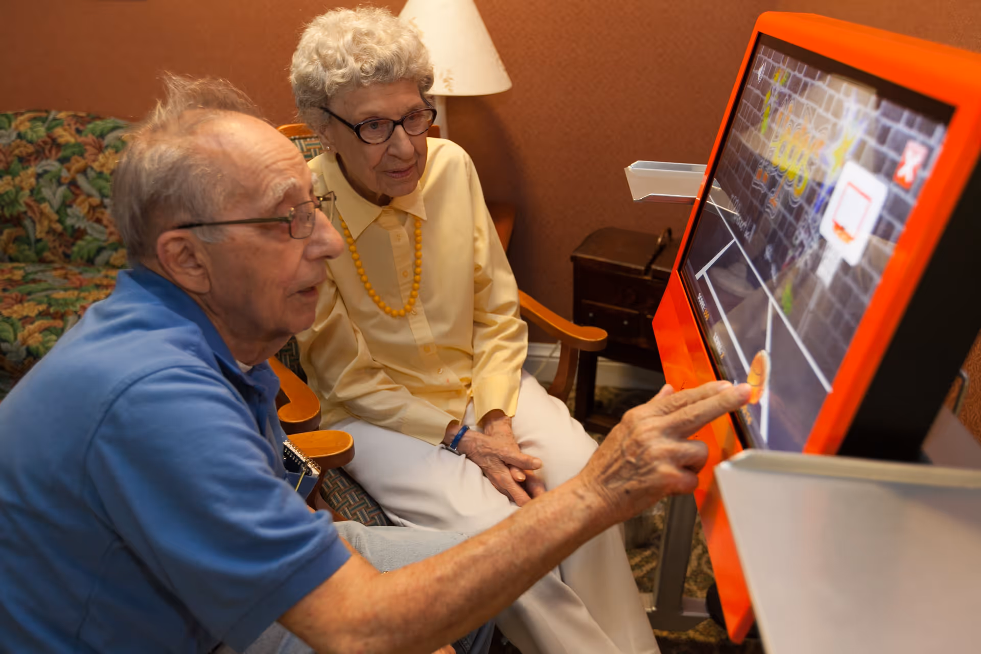 An elderly man and woman sitting in a cozy room with a floral couch and a lamp, interacting with a touchscreen game that displays a basketball hoop and court.