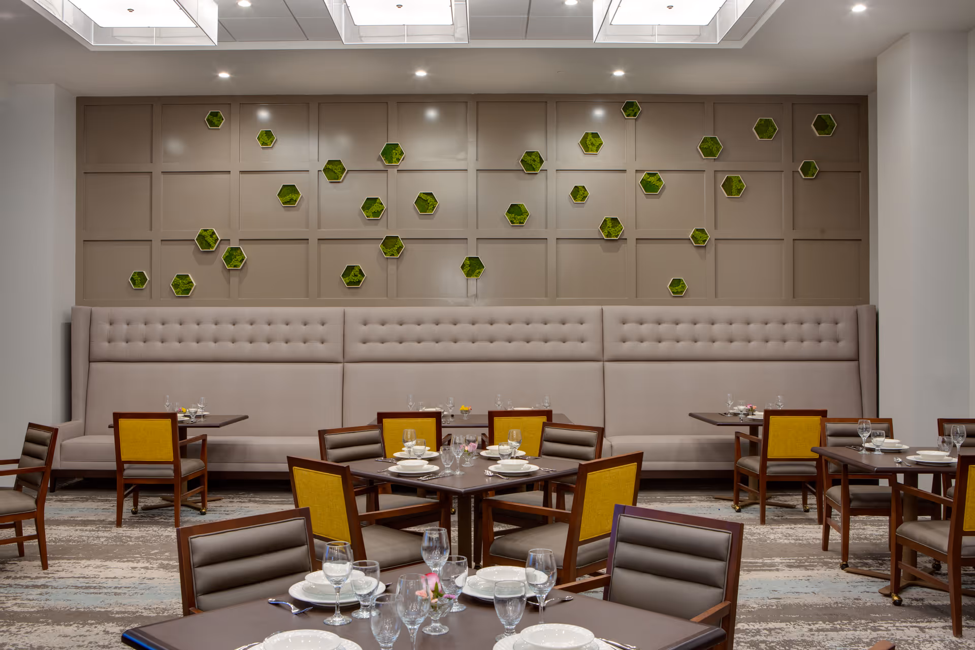 A modern dining room with multiple square wooden tables set with white plates, glasses, and silverware. The chairs have wooden frames with yellow and brown upholstery. A long, tufted beige bench runs along the back wall, which is decorated with small hexagonal green wall accents. The room has a neutral color palette and a patterned carpet.