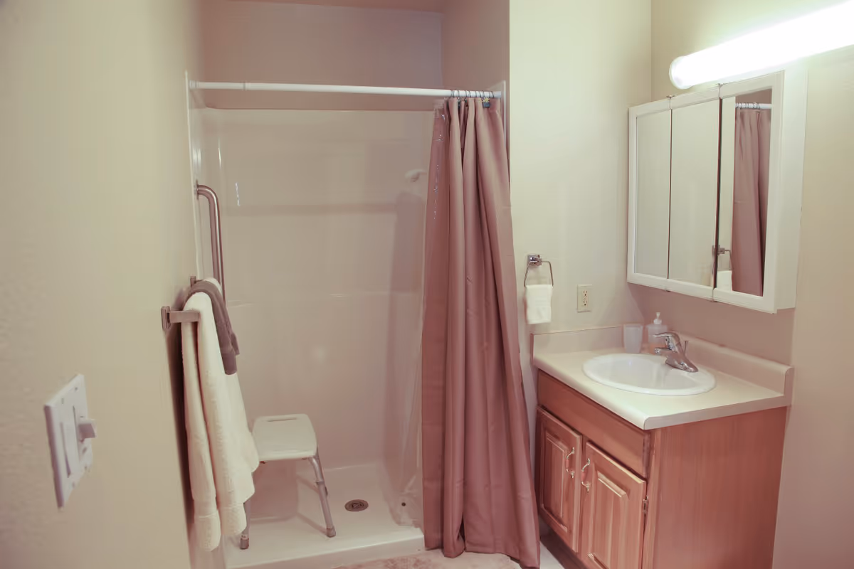 A bathroom with a shower area that includes a white shower chair and a brown shower curtain. There is a towel rack with two towels on the left wall. On the right side, there is a sink with a countertop, a soap dispenser, a cup, and a mirrored medicine cabinet above. The lighting is bright and the walls are light-colored.