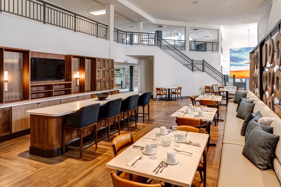 A spacious and modern dining area with multiple tables set with white cups, plates, and silverware. There is a long cushioned bench with pillows along one side and wooden chairs on the other. In the background, there is a large wooden cabinet with a mounted flat-screen TV and a long island with black cushioned bar stools. A staircase with black railings leads to an upper level, and a colorful painting is visible on the far wall.