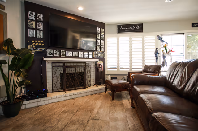 A cozy living room with a large flat-screen TV mounted above a white brick fireplace with a metal screen. The wall around the TV is decorated with black and white photos and movie-themed signs. There is a brown leather sofa and a matching armchair with an ottoman. A large potted plant is on the left side, and windows with white shutters let in natural light. A sign above the windows reads 'Love never fails'.