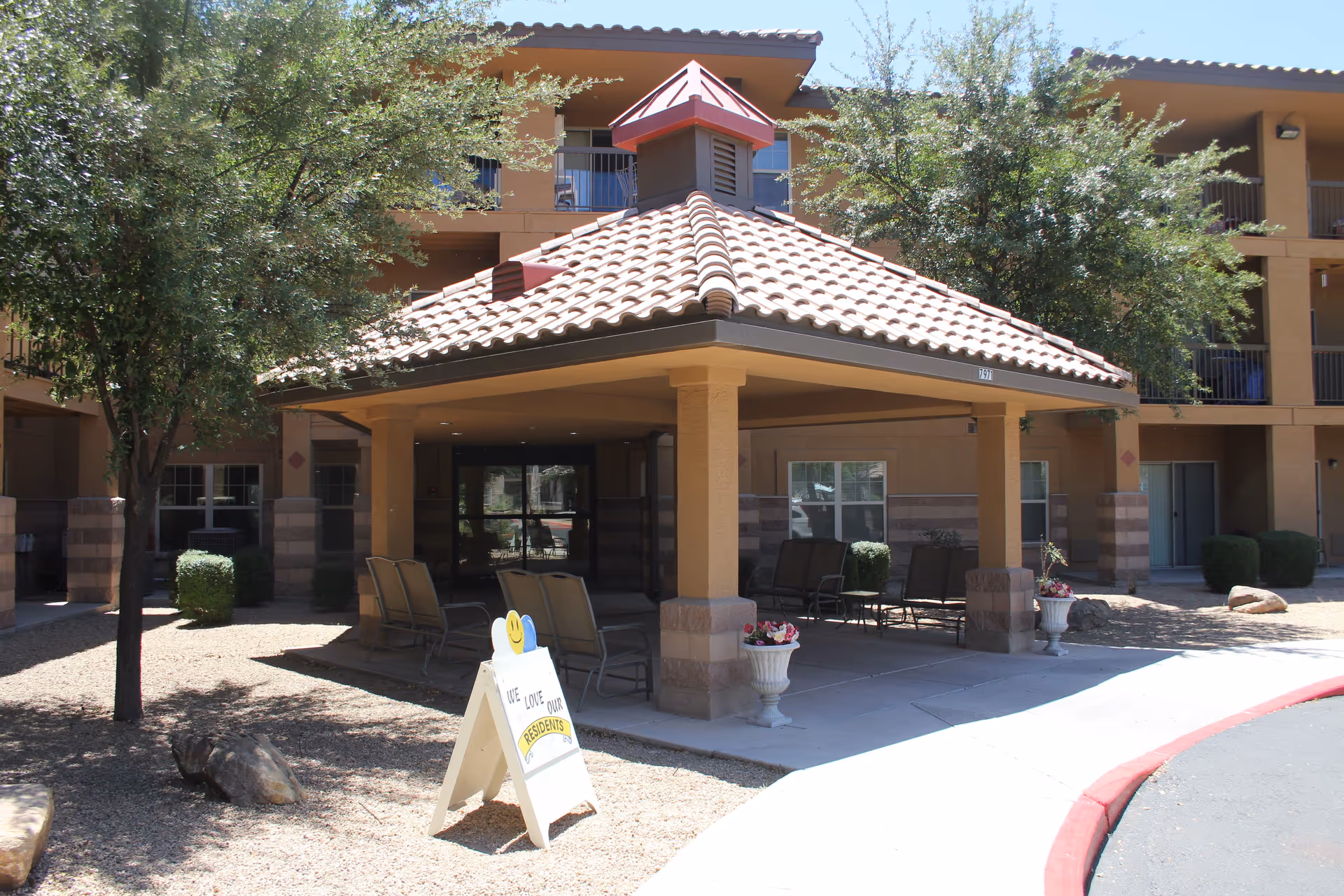 Exterior view of a senior living facility entrance with a covered patio area supported by columns, outdoor seating, trees, and a sign that reads 'We love our residents'. The building has a tiled roof and multiple balconies.