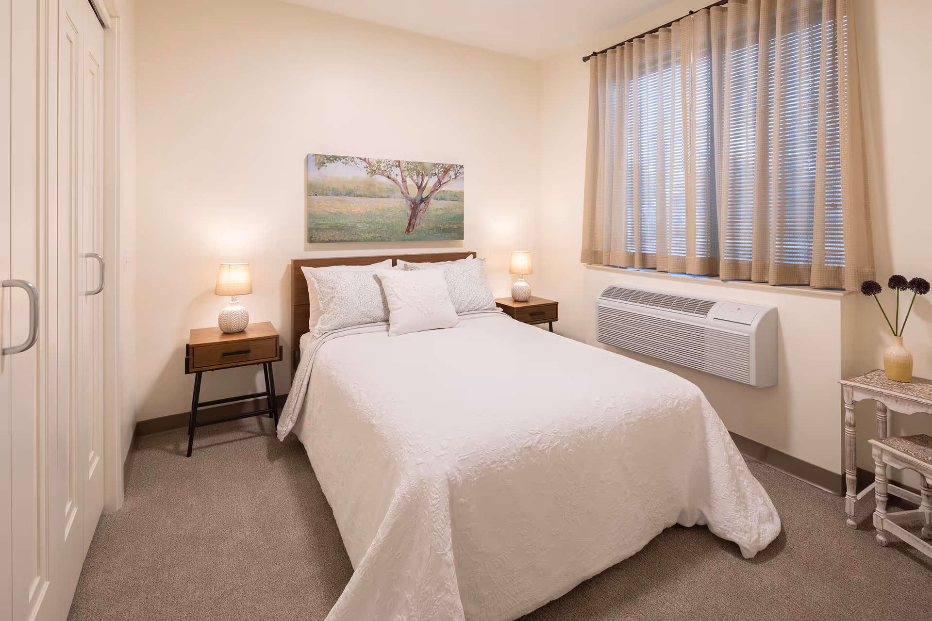 Cozy furnished bedroom with a made bed, two nightstands and lamps, wall art, a window with curtains, and an air conditioning unit.